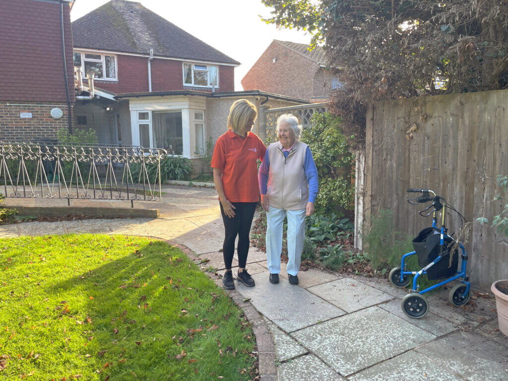 A Beehive Care staff member helping an elderly lady to walk around a sunny garden.